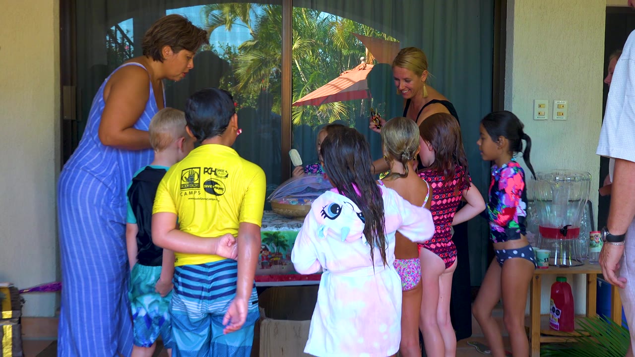 Several children in swimwear stand with their backs and sides to camera while adults supervise near a patio table in San Jose del Cabo, Mexico.