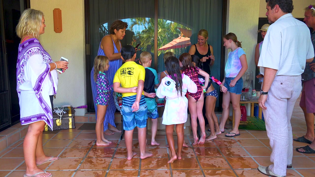 In San Jose del Cabo, children in swimwear cluster on a wet tiled patio while adults stand around them near reflective sliding glass doors.