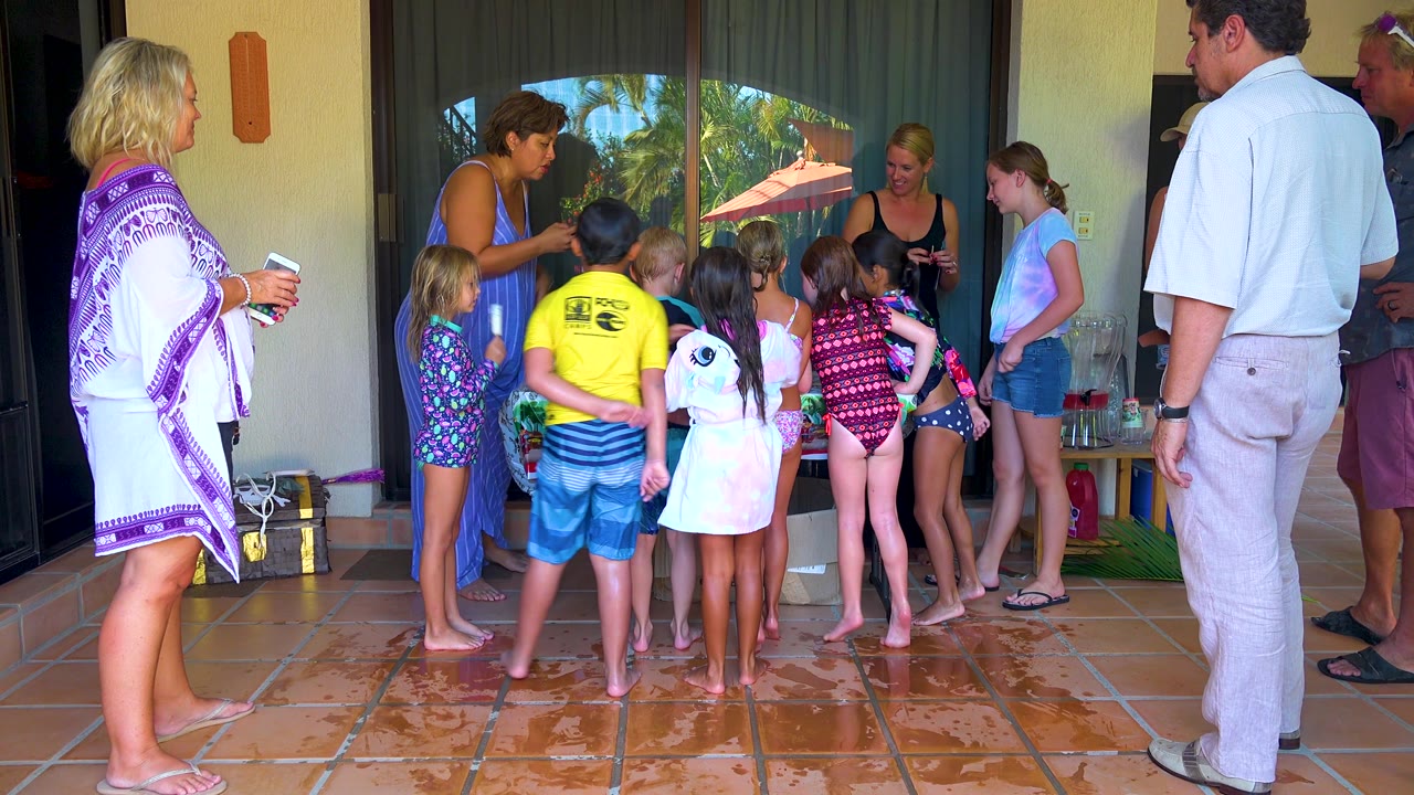 The patio group in San Jose del Cabo shifts slightly, with children in colorful swimwear centered and adults standing at the sides near cups and towels.