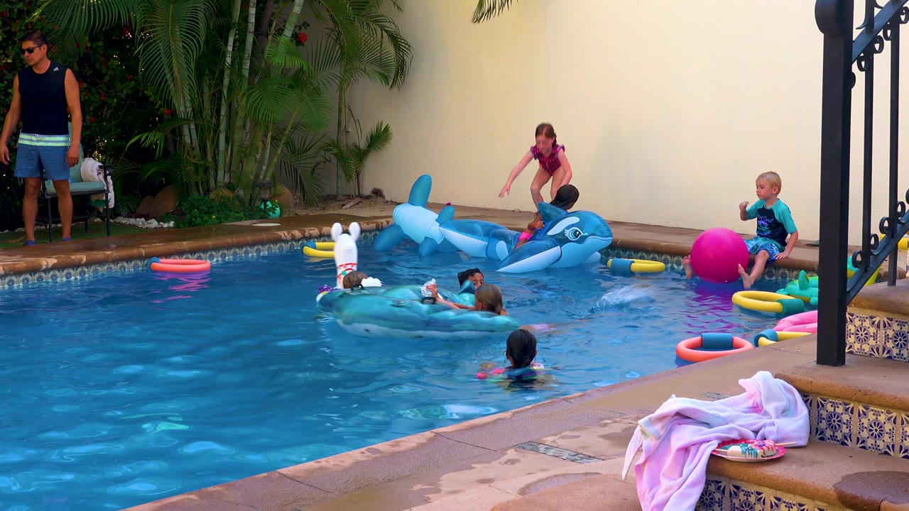 Children in San Jose Del Cabo play in a backyard swimming pool with a blue dolphin float, white animal float, colorful rings, a pink ball, towels on steps, and an adult standing at left.