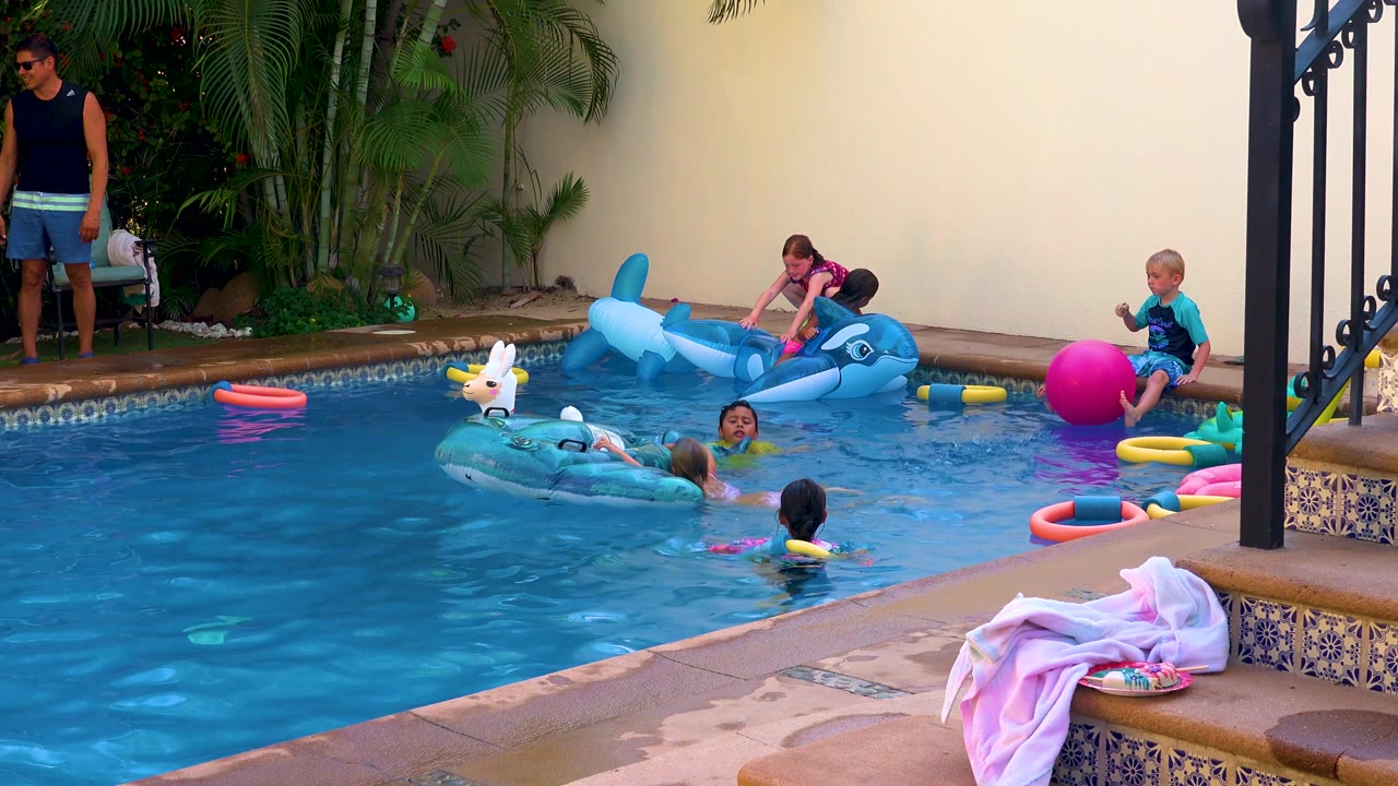 At 0:02, the San Jose Del Cabo pool scene shows a child near the bright pink ball on the right while others move around the white animal float and blue dolphin float in the water.