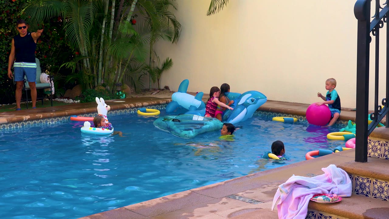 Children float and swim around dolphin and swan inflatables in a backyard swimming pool.