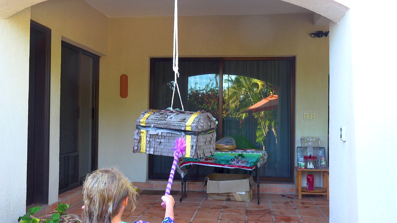 At 0:00 in San Jose Del Cabo, a child seen from behind raises a purple striped stick toward a gray hanging pinata on a tiled patio.