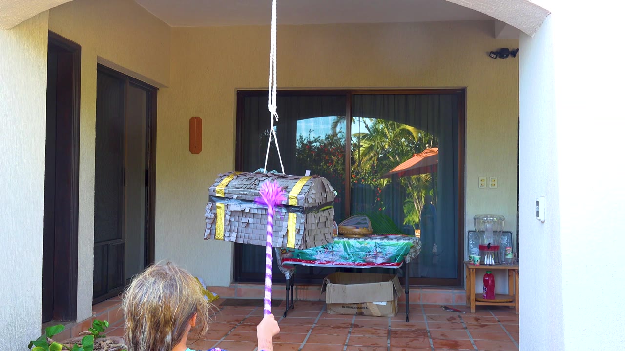 At 0:02, the purple striped stick reaches the front of the gray pinata as the child strikes upward from the foreground on the San Jose Del Cabo patio.