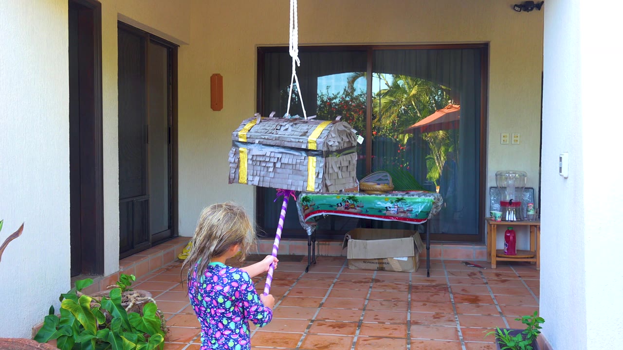 In San Jose Del Cabo, a young girl faces a hanging treasure-chest piñata while holding a striped stick on a terracotta-tiled outdoor patio.