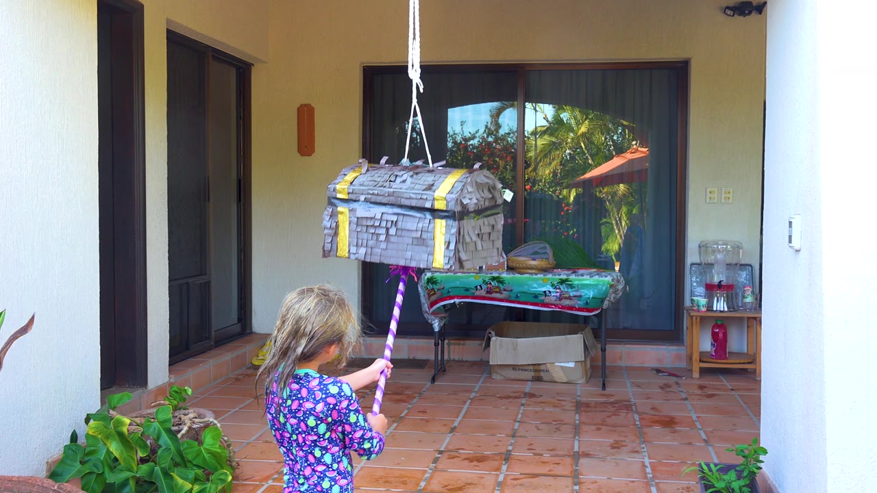 The girl remains below the hanging piñata in San Jose Del Cabo, with the stick angled upward and the tiled patio and glass doors behind her.