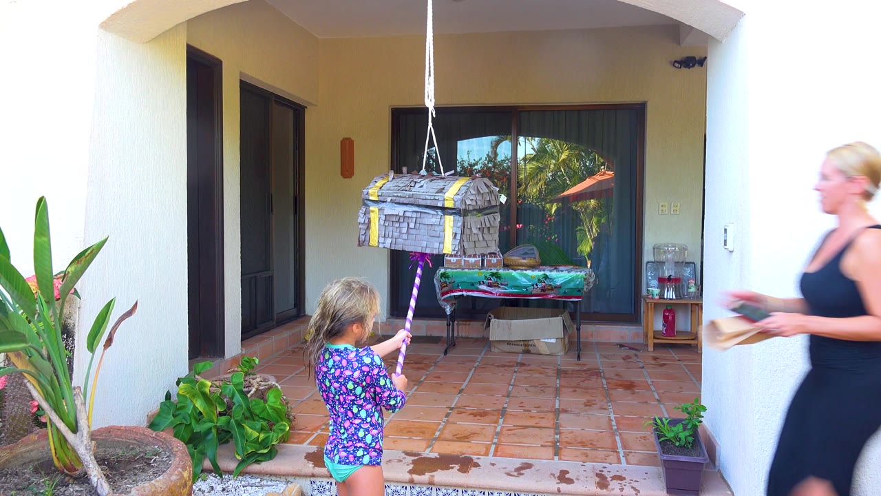 A woman in a black dress enters from frame right carrying a book while the girl faces the hanging piñata on the San Jose Del Cabo patio.