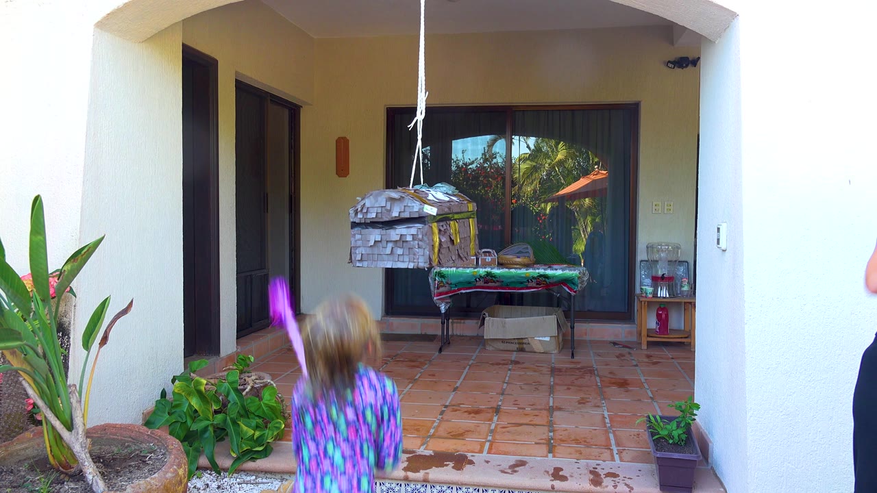 At 0:00 in San Jose del Cabo, a child stands in the foreground with a purple stick aimed toward a hanging wooden piñata on a terracotta tiled patio.