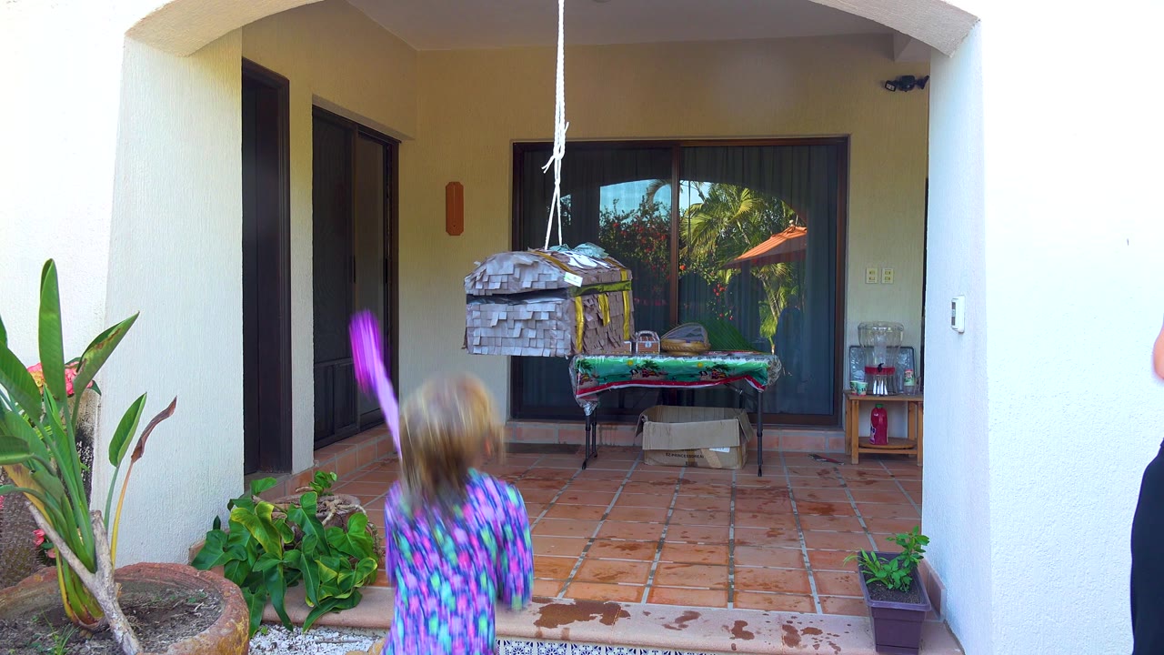 At 0:01 in San Jose del Cabo, the child swings the purple stick upward toward the hanging piñata while plants, glass doors, and a covered table remain in the background.
