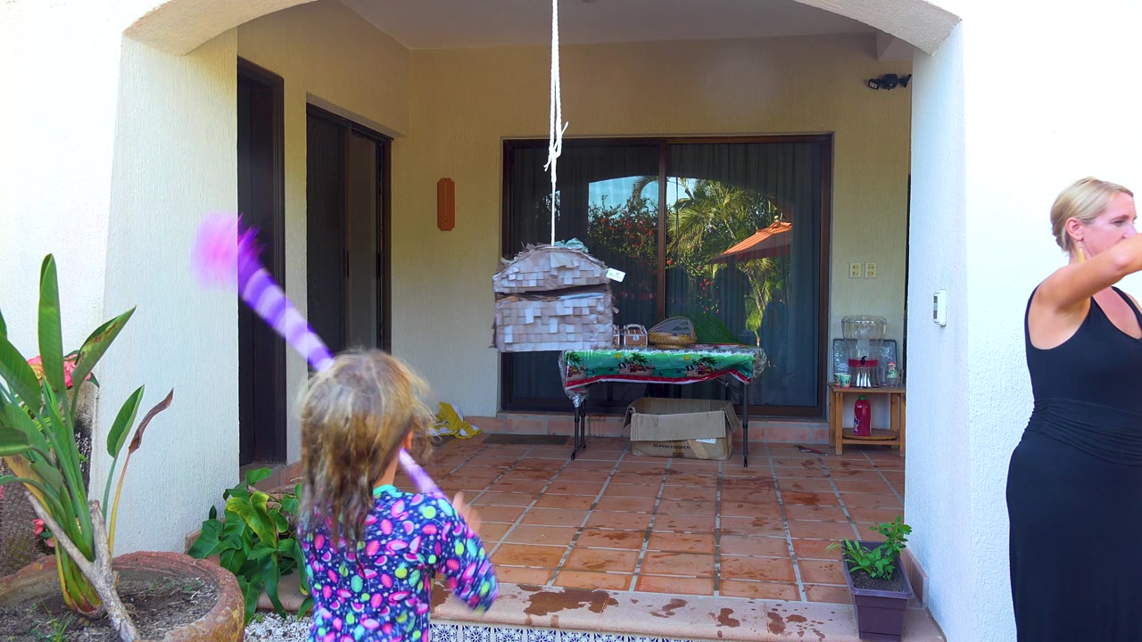 At 0:02 in San Jose del Cabo, the child raises the purple stick near the piñata while an adult woman is partly visible at frame right on the patio.