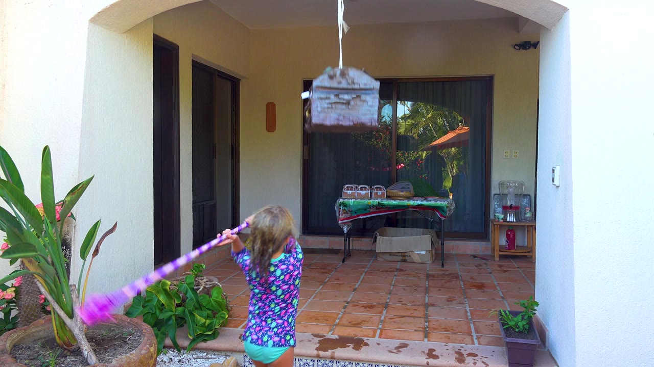 In San Jose Del Cabo, a child in a colorful swim shirt prepares to swing a purple stick at a hanging treasure-chest piñata on a covered tiled patio.