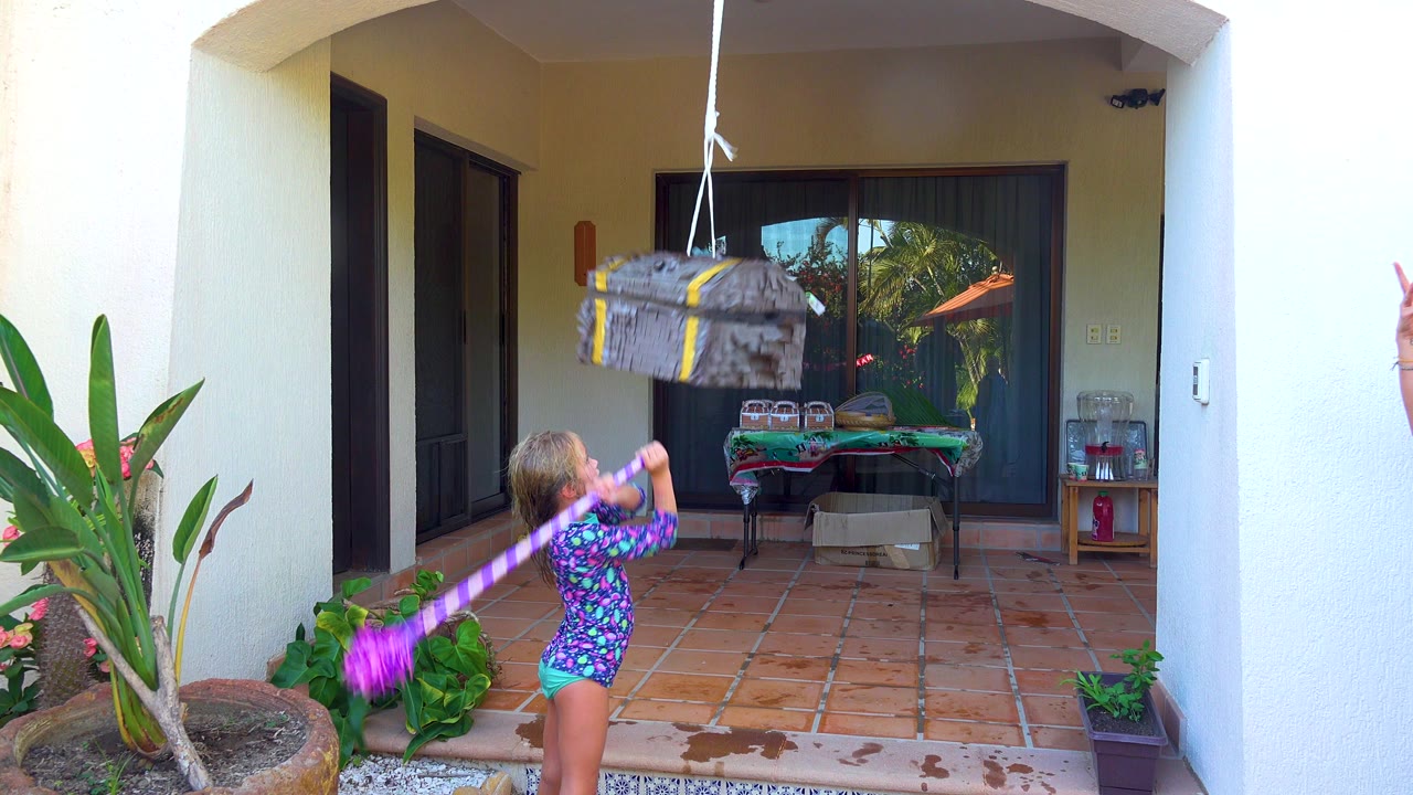 In San Jose Del Cabo, a child in a patterned swimsuit swings a purple stick from the left side of a tiled patio toward a gray piñata hanging near the top of frame.