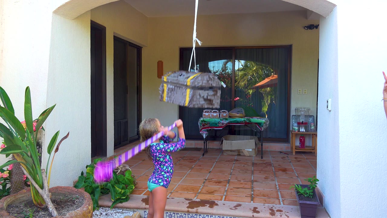 Child continues swinging at a hanging piñata beside potted plants.