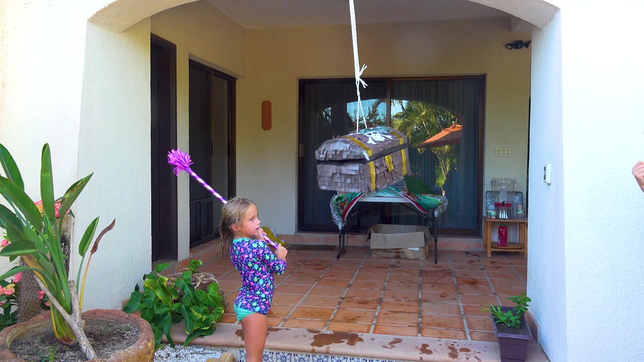 In San Jose Del Cabo, the child faces the hanging piñata with the purple stick lifted, with a large potted plant at left and reflections in the dark sliding glass doors behind.