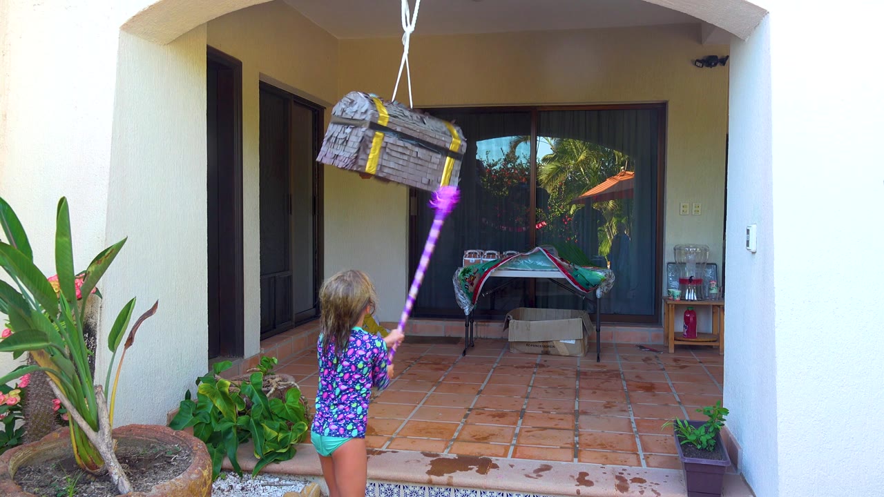 Child continues swinging at the piñata beneath a covered patio.
