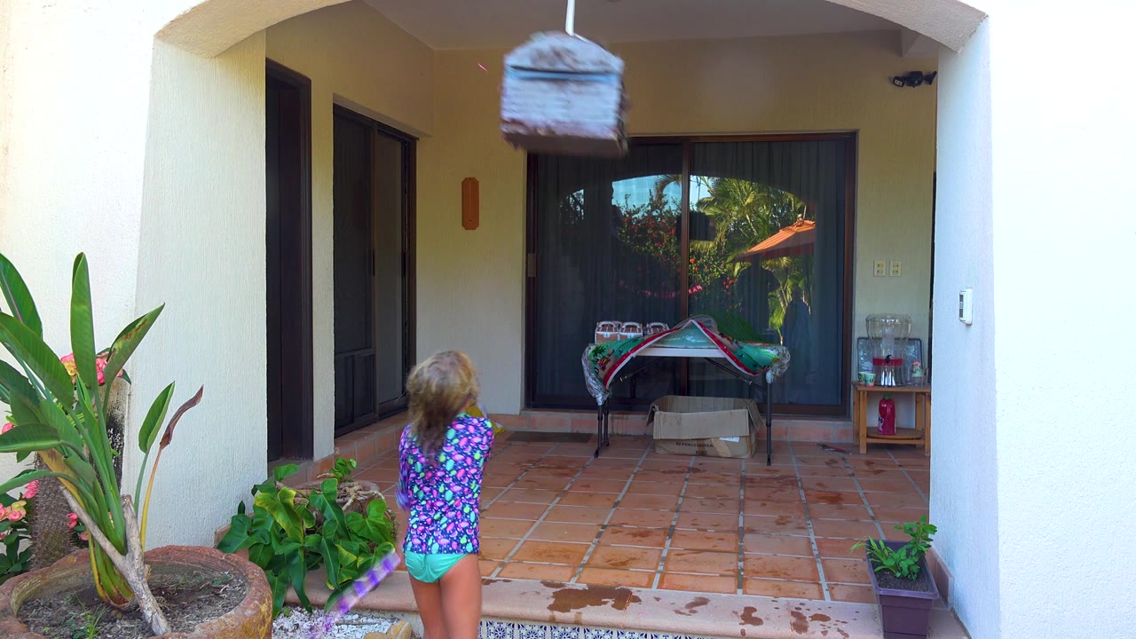 At 0:02 in San Jose Del Cabo, the piñata swings upward near the top of the frame as the child lowers the purple stick on the tiled patio below.