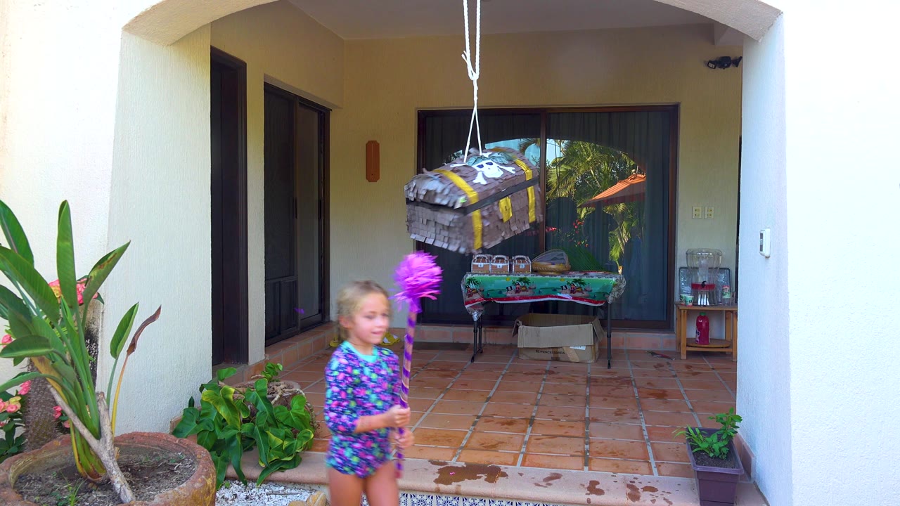 A child in a purple floral outfit stands on a tiled patio in San Jose Del Cabo, holding a purple stick as a hanging piñata faces the camera.