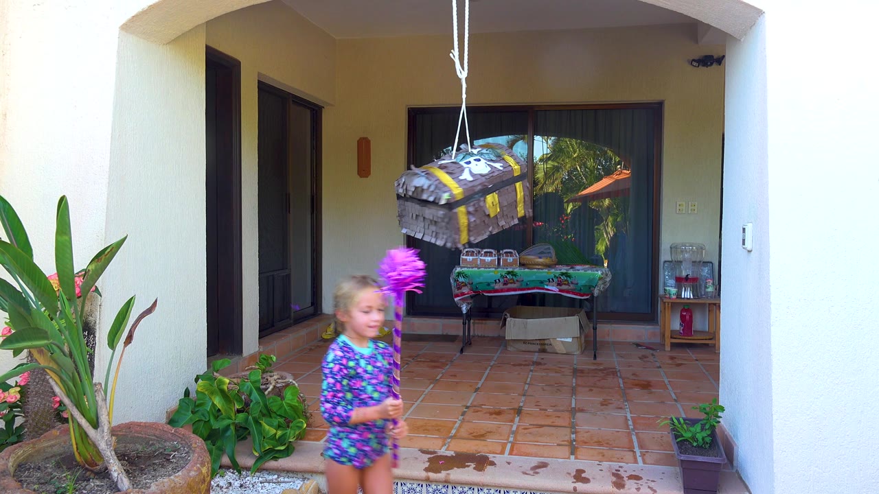 Child steps across the patio with the purple stick beside the hanging piñata.