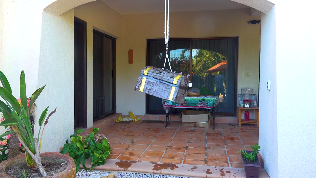 A hanging piñata is centered over a terracotta tiled patio in San Jose del Cabo, with potted plants, a table, and sliding doors behind it, and no visible people.