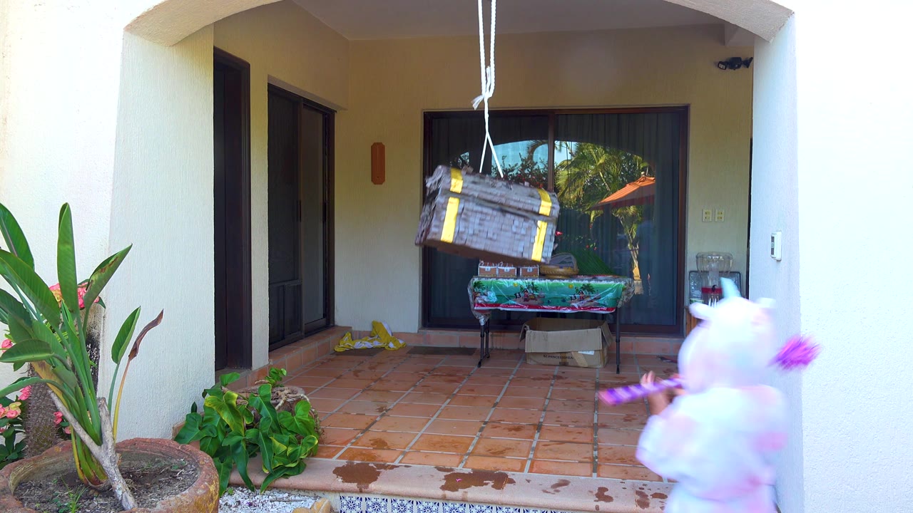 A child in a white bunny costume enters from the right edge of the San Jose del Cabo patio while holding a purple striped stick below the hanging piñata.