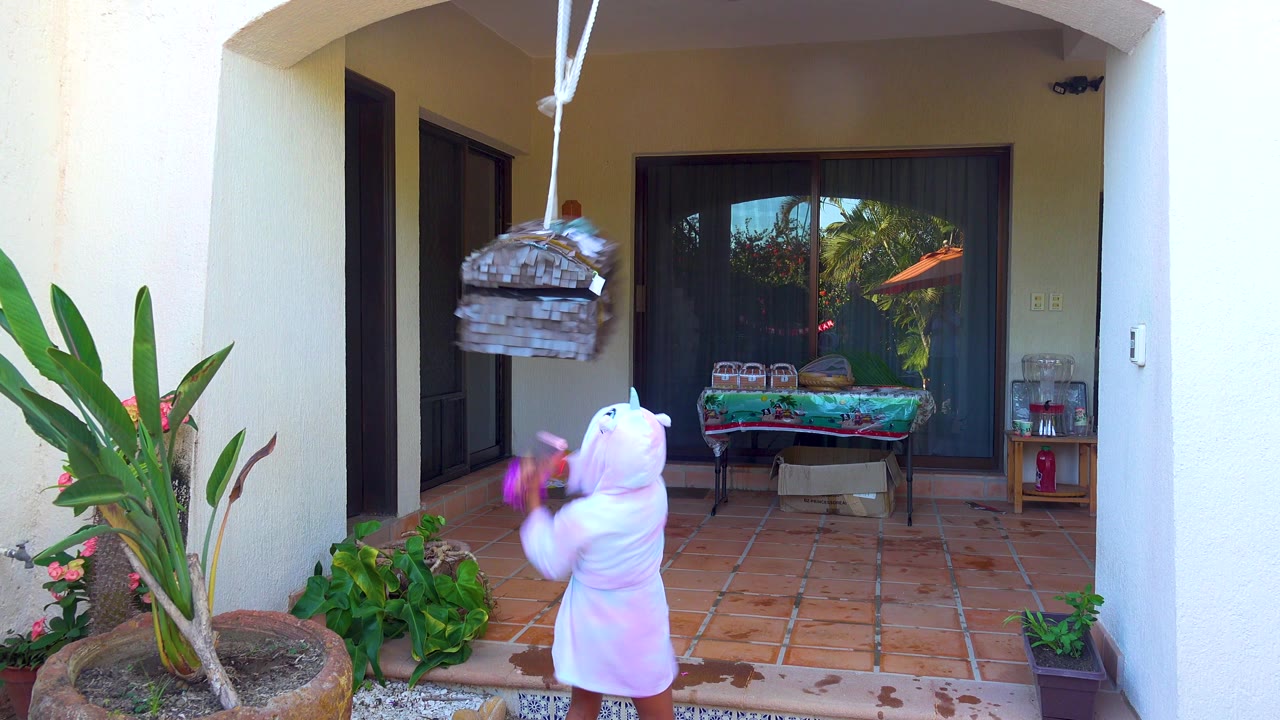 In San Jose Del Cabo, a child in a white hooded onesie stands on a terracotta tiled patio and swings a purple stick toward a hanging piñata near potted plants and sliding glass doors.