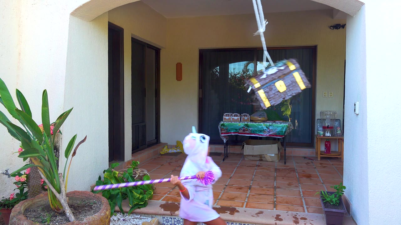 In San Jose Del Cabo, the chest-shaped piñata swings high to the right while the child in the white onesie holds the purple stick across the patio foreground.