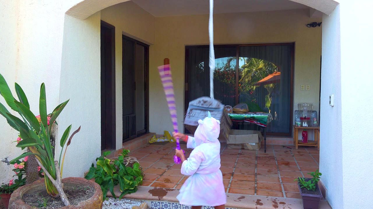 At 0:00 in San Jose del Cabo, a child in a white hooded outfit stands below a hanging piñata on a terracotta tiled patio with potted plants and glass doors behind.