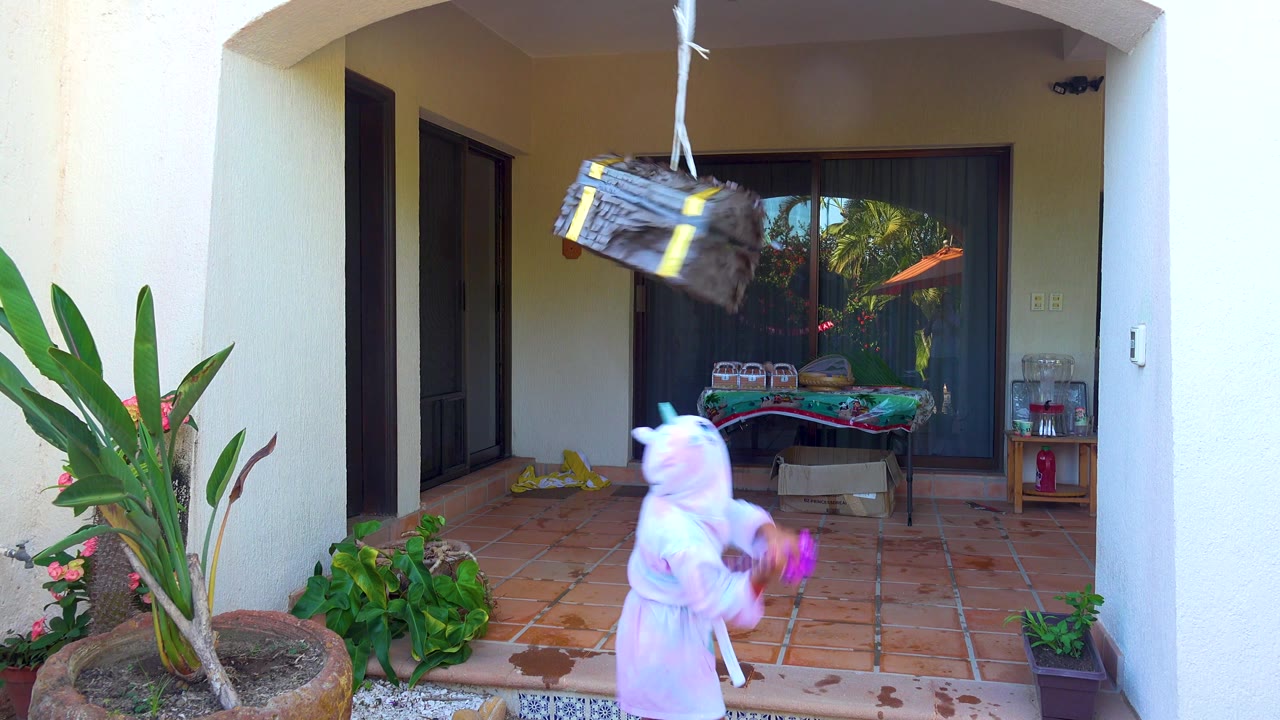 At 0:02 in San Jose del Cabo, the treasure-chest piñata swings high above the child, who holds the purple stick near the right side of the patio frame.