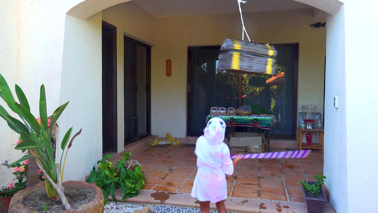 In San Jose del Cabo, a child in a white hooded robe faces a hanging chest-shaped piñata on a tiled outdoor patio with potted plants and sliding glass doors behind.