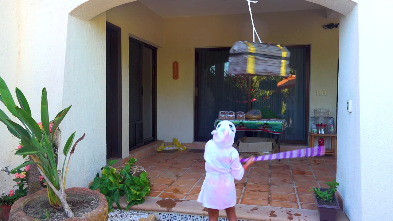 Child holds a purple striped stick toward a swinging piñata on the patio.
