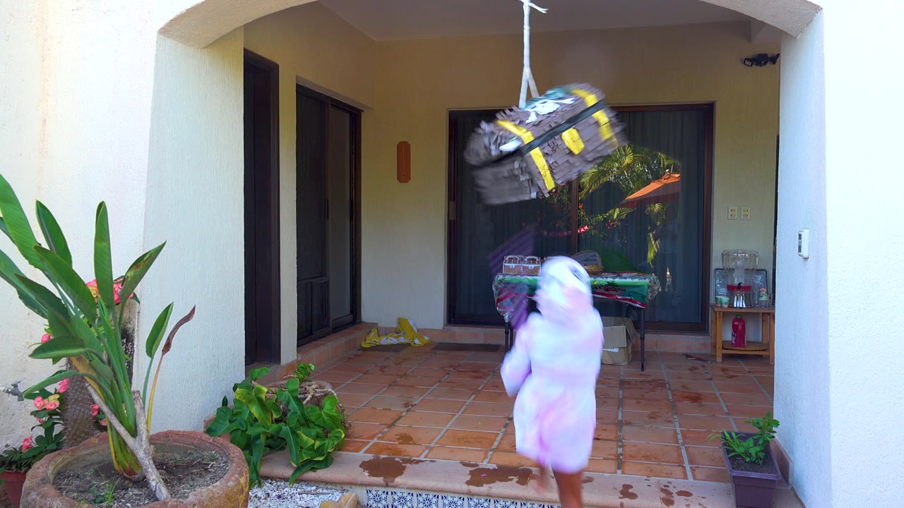 In San Jose del Cabo, the piñata swings high to the right while the child in a white hooded robe turns below it holding the purple striped stick on the patio.