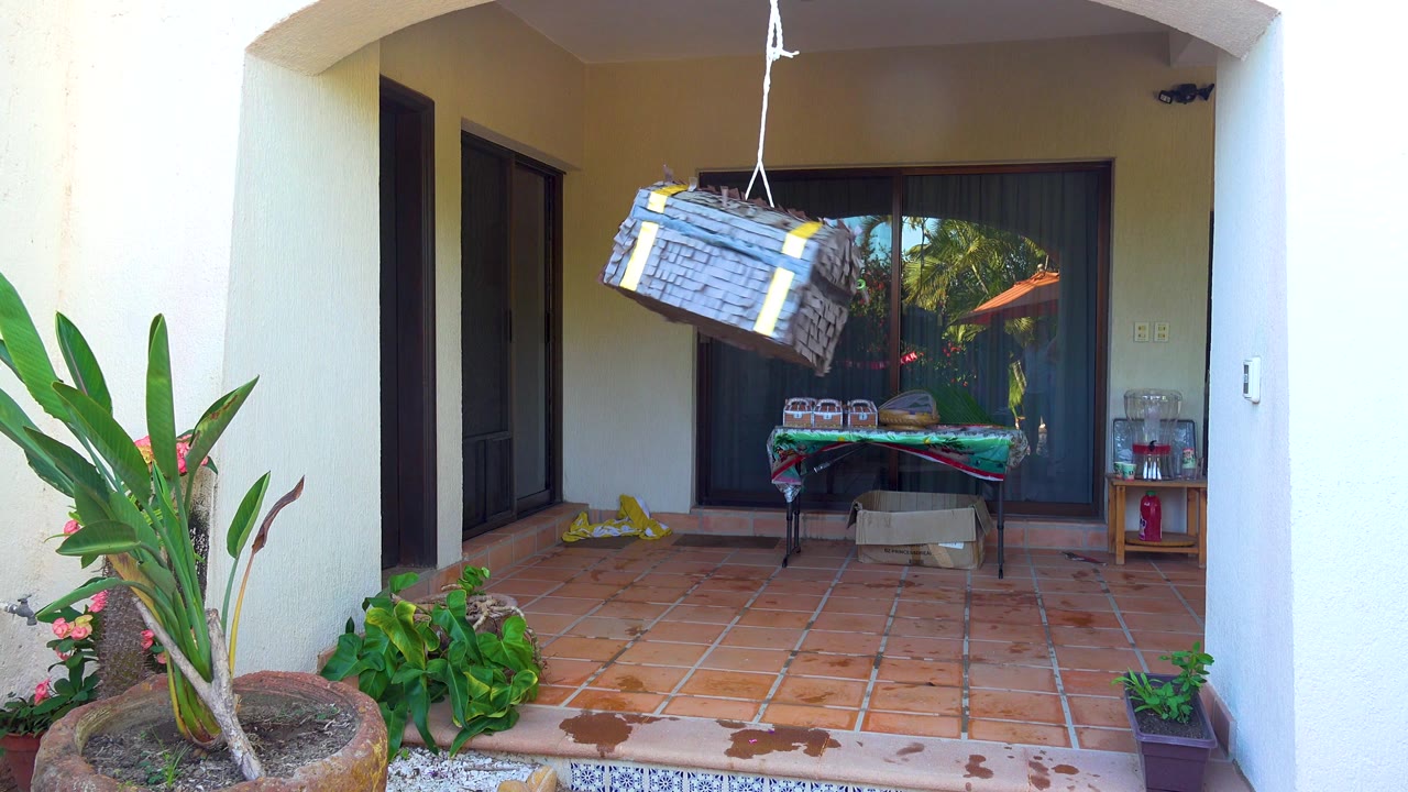 A gray hanging piñata swings near the upper center of a tiled outdoor patio in San Jose Del Cabo, with potted plants at left, a table by sliding glass doors, and no visible people.