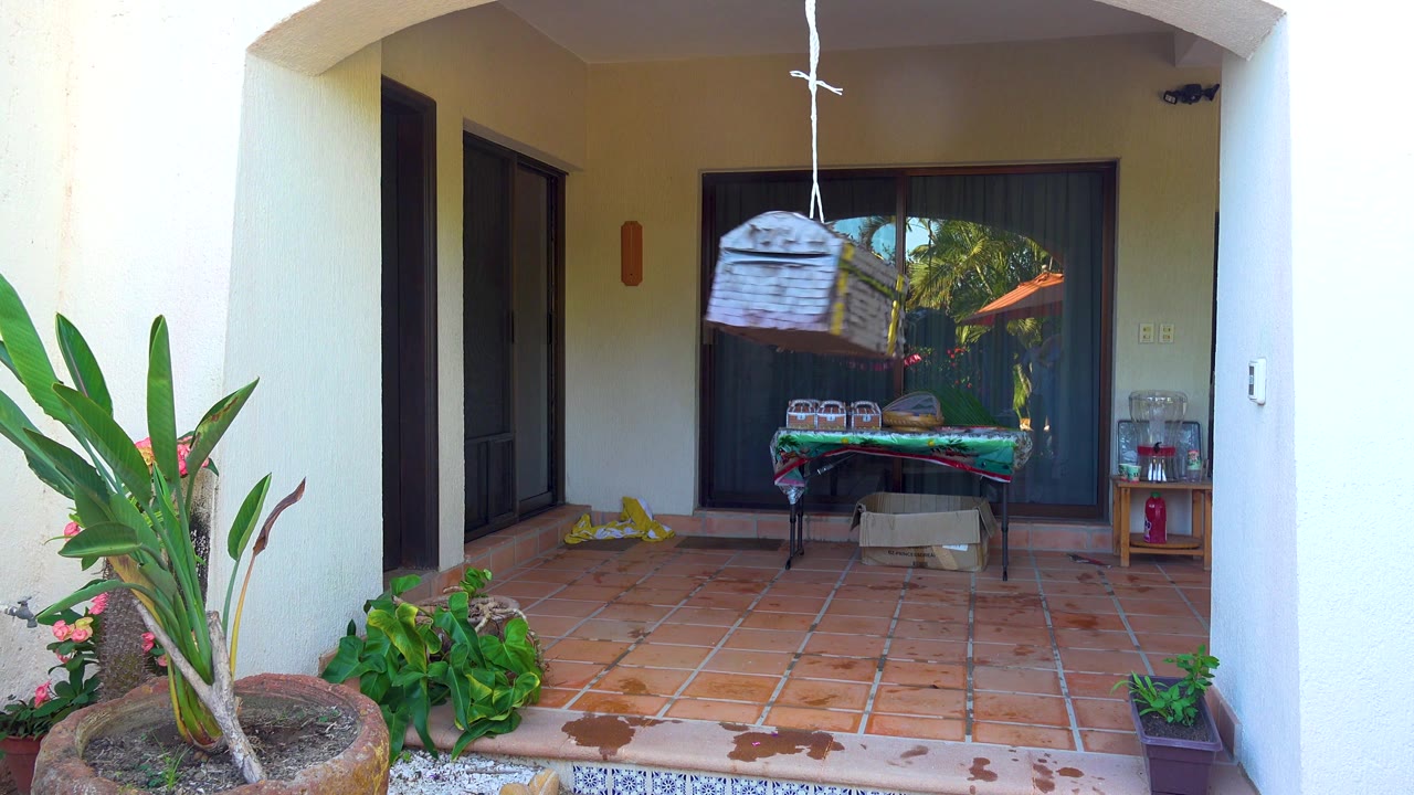 The hanging piñata shifts toward the center of the shaded patio in San Jose Del Cabo, above a table and cardboard box, with plants and tiled flooring visible and no visible people.