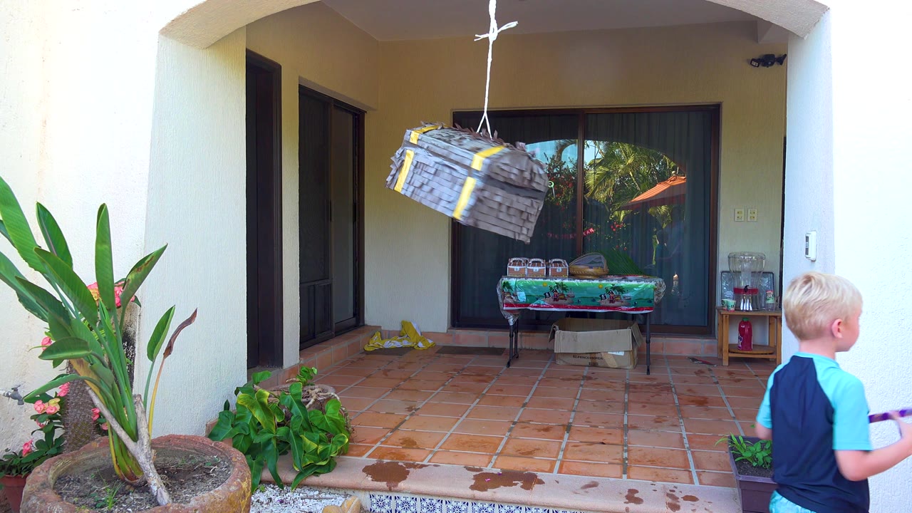 The child is partly at the right edge of the San Jose Del Cabo patio frame while the suspended gray pinata tilts, with green plants and glass doors behind it.