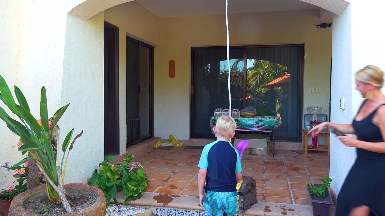 A woman in a black dress steps in from the right near the child and purple piñata on the San Jose del Cabo patio, holding an object in one hand.