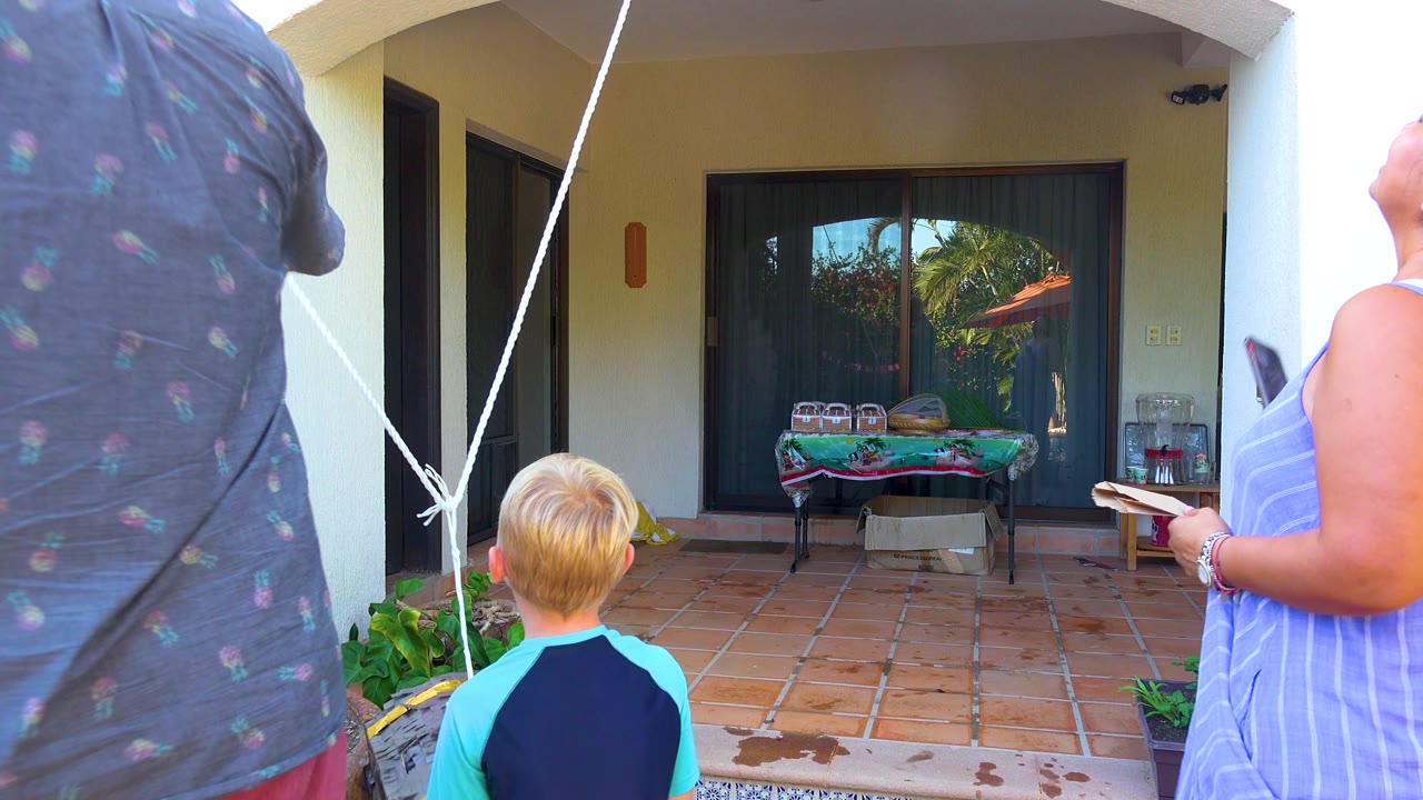 The man stands with his back partly to camera near the diagonal white rope, while the woman at right holds a plate and phone and the child faces the shaded patio.