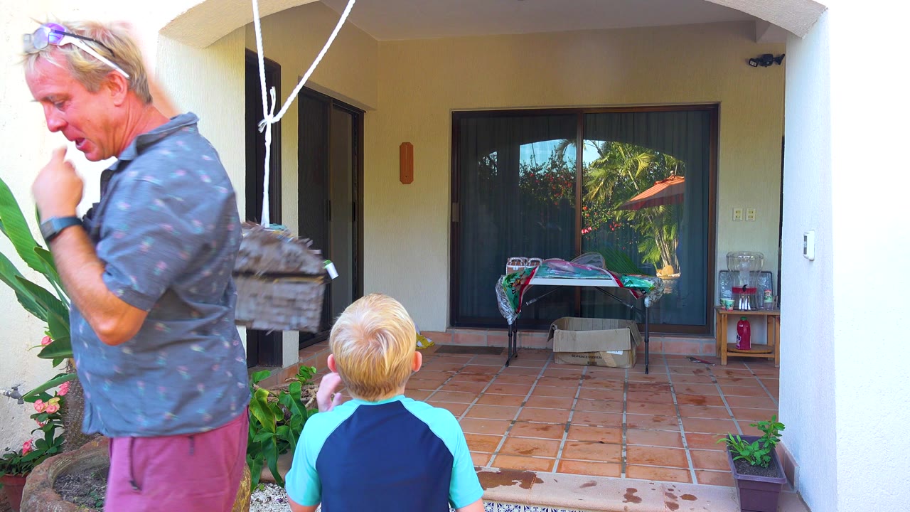 In San Jose Del Cabo, Baja California Sur, an adult man stands at left while a blond child faces a small hanging piñata on a tiled patio with plants and sliding doors behind them.