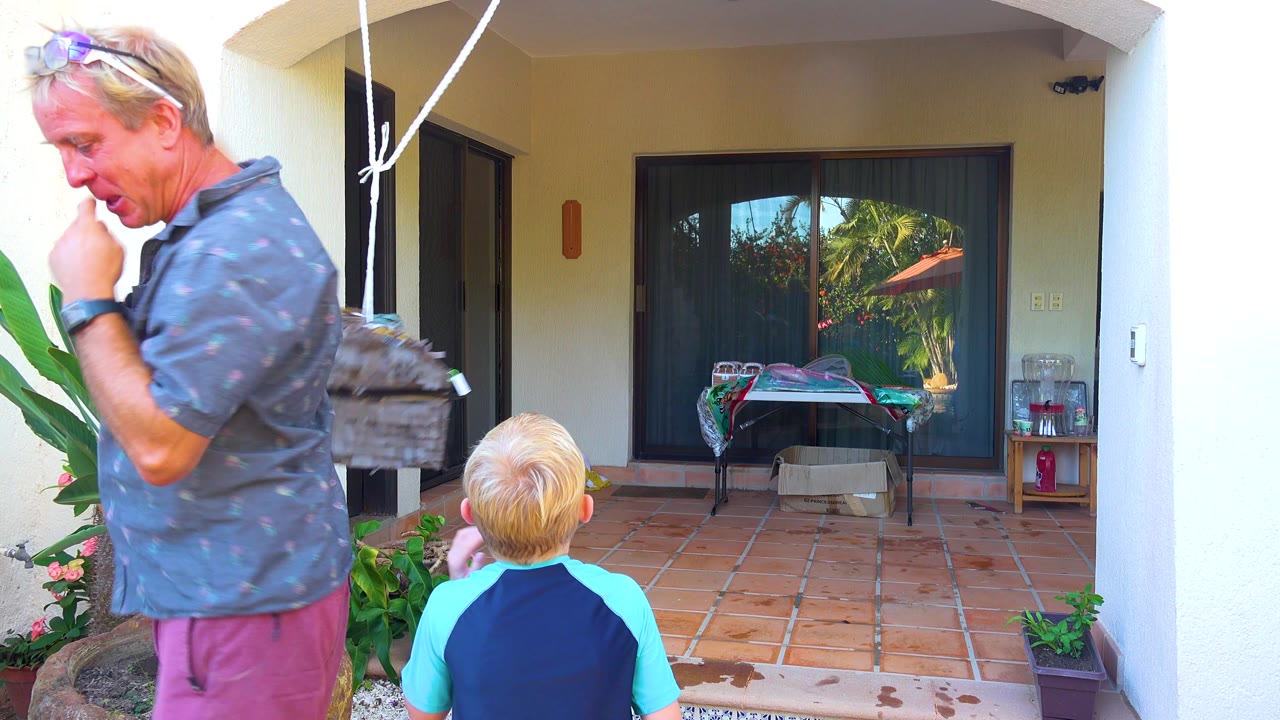 Adult man and child remain near a hanging piñata on a shaded tiled patio.