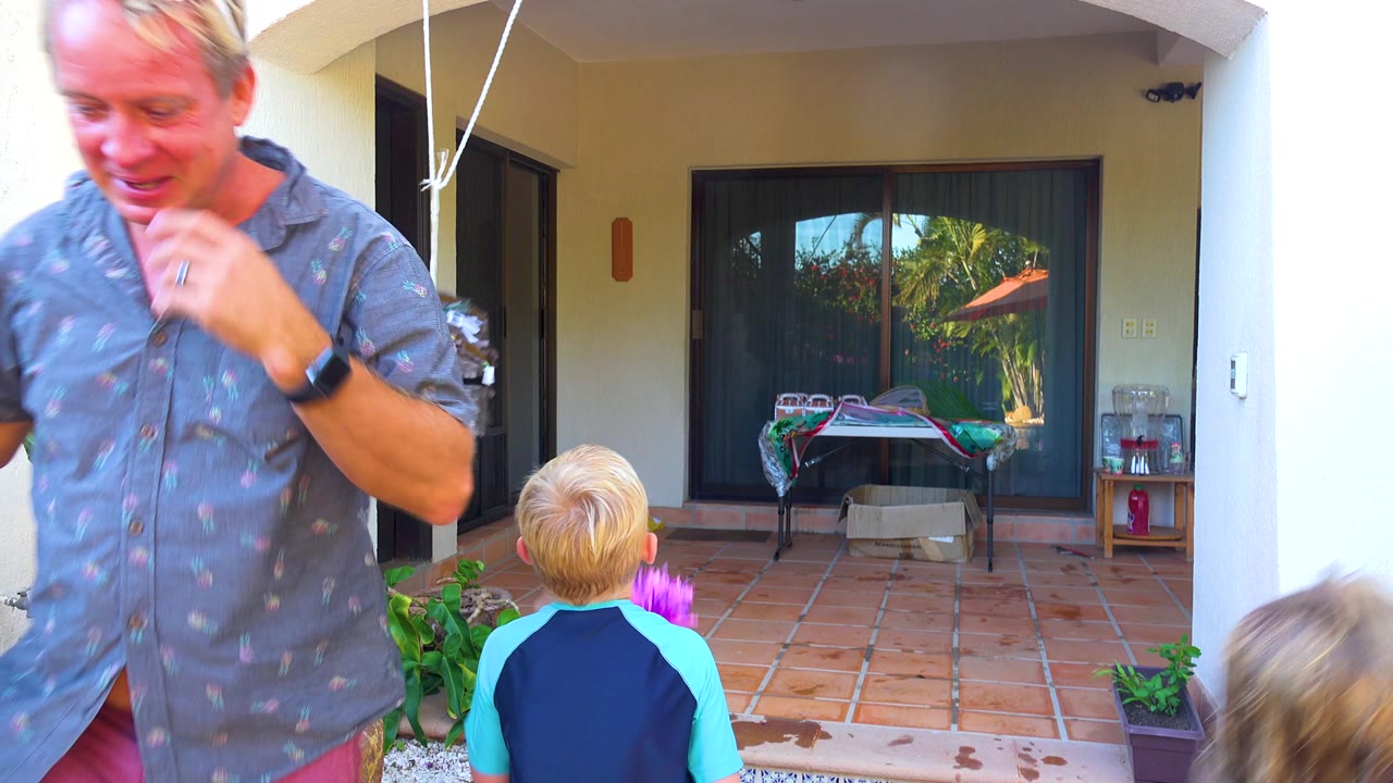 An adult man moves close to the camera with his face visible while the child remains centered below the hanging piñata on the San Jose Del Cabo patio.
