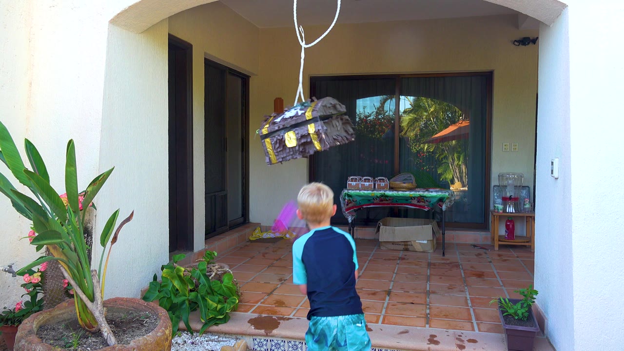 In San Jose Del Cabo, Baja California Sur, a child in a blue shirt and green shorts stands with a purple stick beneath a hanging piñata on a tiled patio.