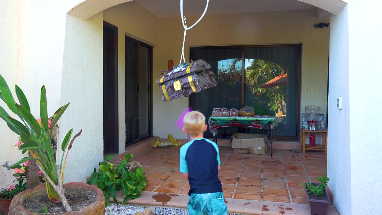 In San Jose Del Cabo, Baja California Sur, the child raises a purple stick toward the hanging piñata while standing on the terracotta patio near potted plants.