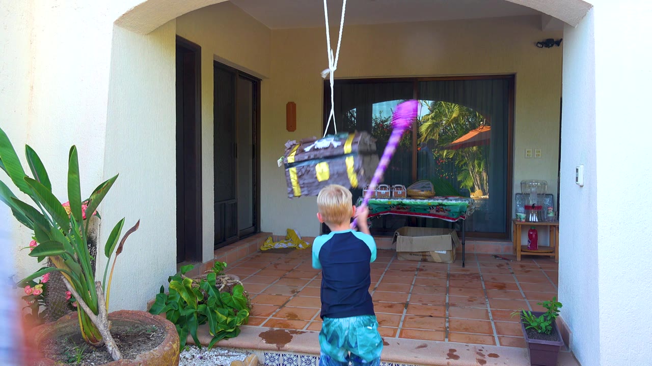 In San Jose Del Cabo, Baja California Sur, the child swings the purple stick up toward the treasure-chest piñata as it hangs near the patio roof opening.