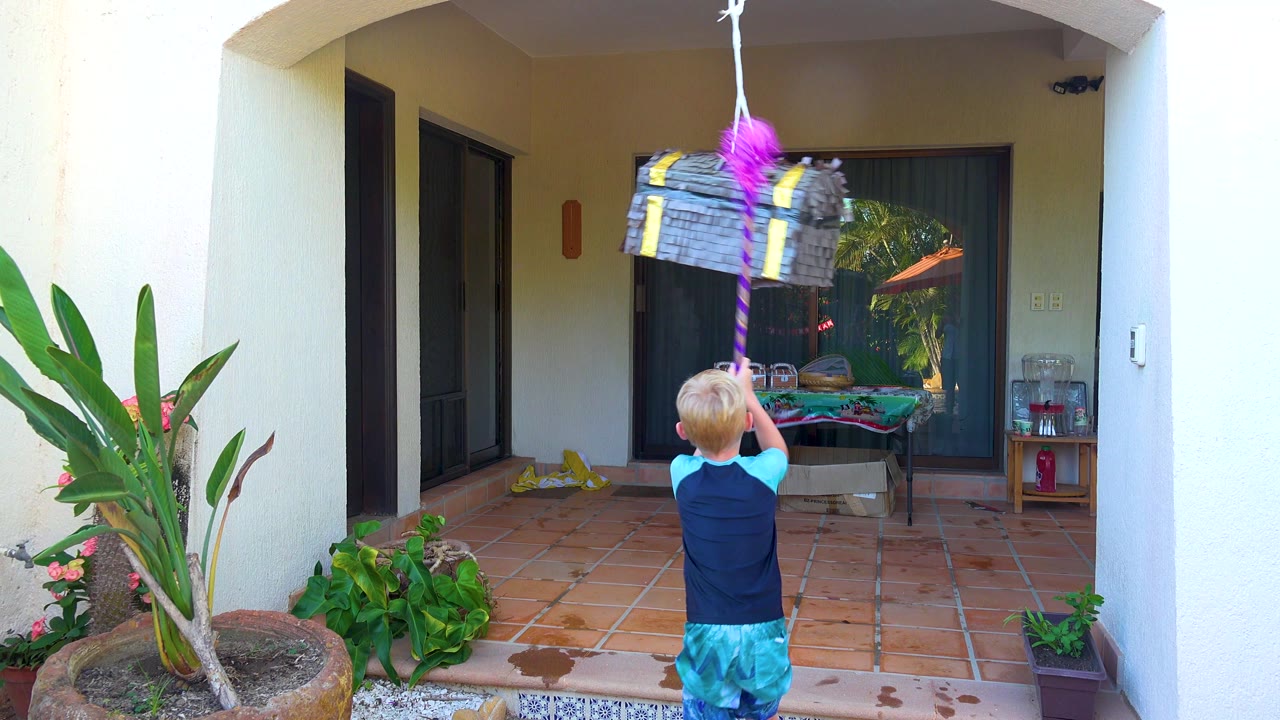 In San Jose Del Cabo, a blond child seen from behind raises a pink stick toward a hanging piñata on a shaded tiled courtyard patio.