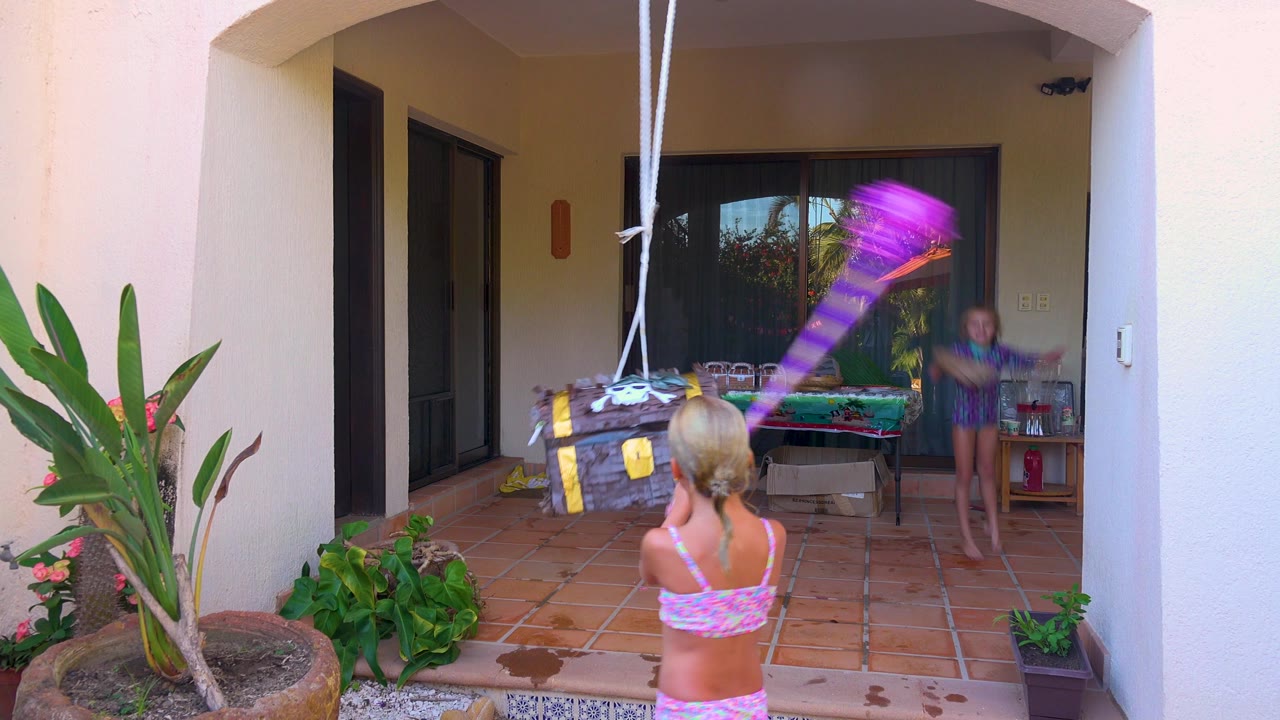 In San Jose Del Cabo, Mexico, a child in a pink swimsuit stands with her back to camera as a hanging treasure-chest piñata swings in front of a shaded patio and sliding glass doors.