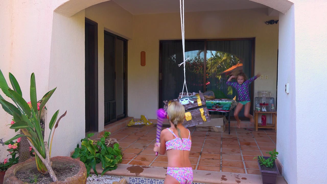 A child in a pink swimsuit holds the purple stick upright while the piñata hangs lower in front of a tiled patio and a second child stands by the doorway in San Jose Del Cabo, Mexico.