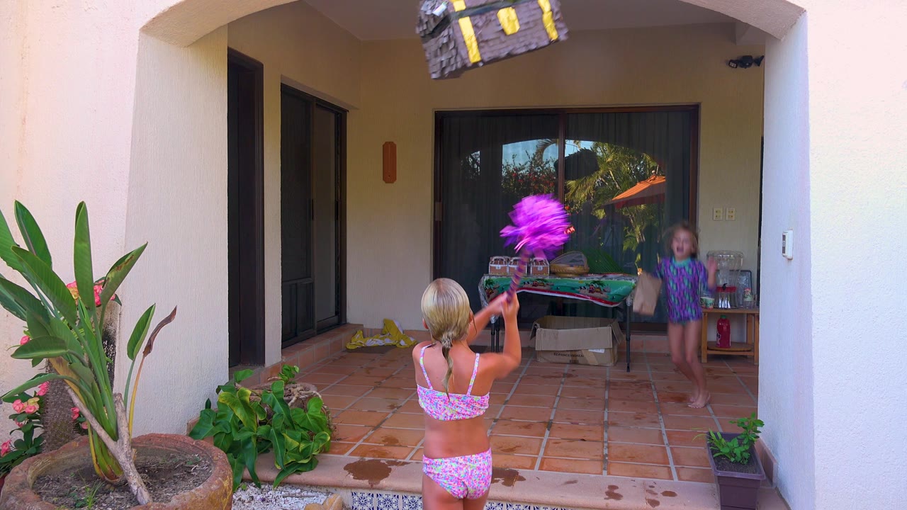 In San Jose Del Cabo, a child in a pink swimsuit stands with a stick aimed toward a hanging treasure chest piñata while another child watches near a patio table.