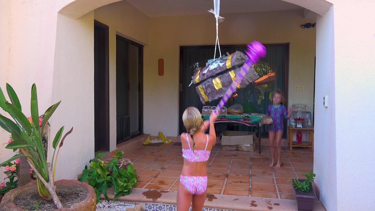 The treasure chest piñata swings high above the child in the pink swimsuit while the second child watches from beside the patio table in San Jose Del Cabo.