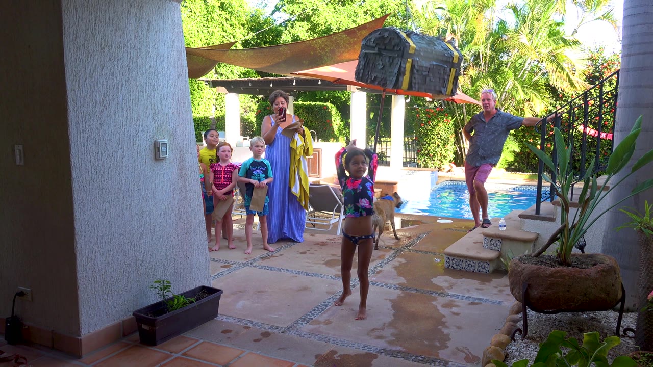 In San Jose Del Cabo, the child raises the large black toy above their head while children and adults watch from the poolside patio under umbrellas and garden shade.