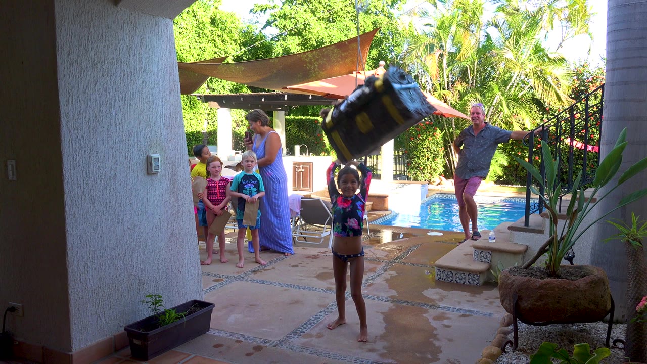 At 0:00 in San Jose Del Cabo, a child stands on wet patio tiles swinging a large dark bag, with children, adults, a swimming pool, stairs, umbrellas, and greenery behind her.