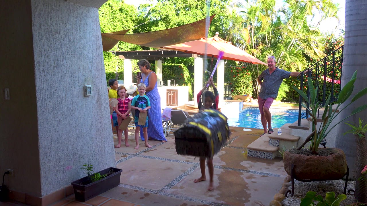 At 0:02, the large bag crosses the foreground in motion blur while the child stands beneath it, with the pool, umbrellas, steps, adults, children, and garden plants in the background.